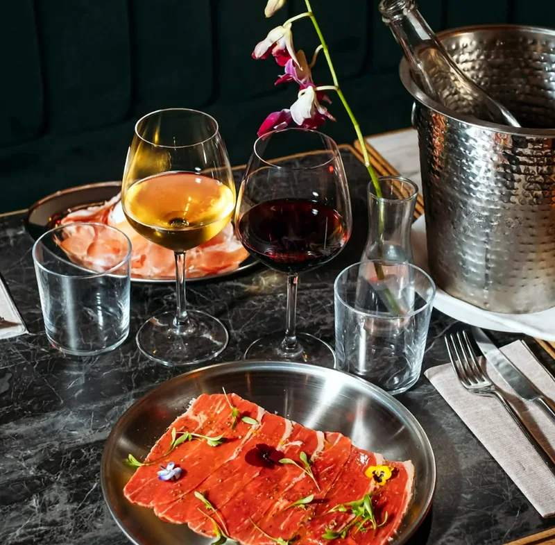Il tavolo da Biffi: marmo scuro, calici di bianco e di rosso, carpaccio guarnito con fiori eduli e secchiello del ghiaccio