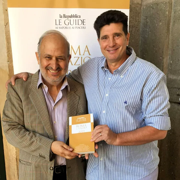 Due persone sorridenti con una guida gastronomica de la Repubblica, davanti al pannello dell'evento