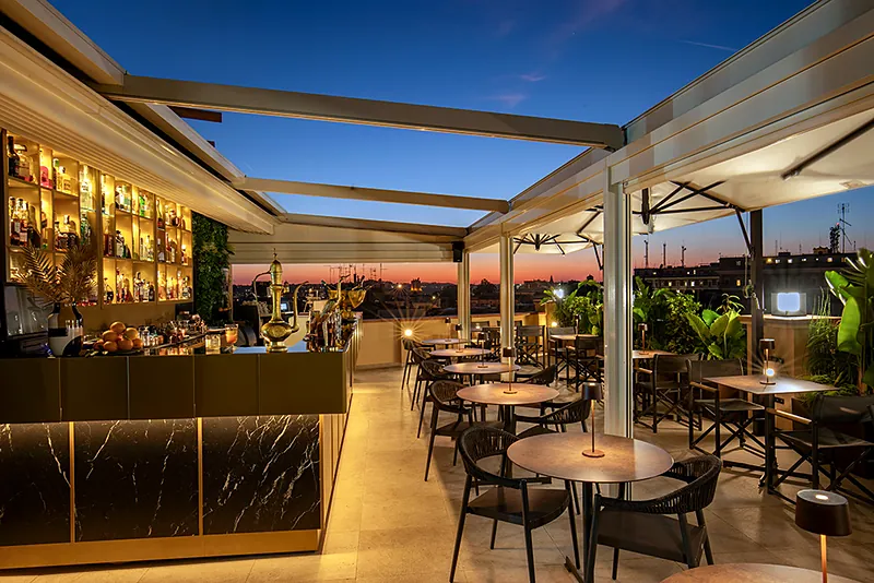 Il bancone del bar e i tavoli sotto la pergola di Masa Roof Top, con il cielo del tramonto sui tetti di Roma
