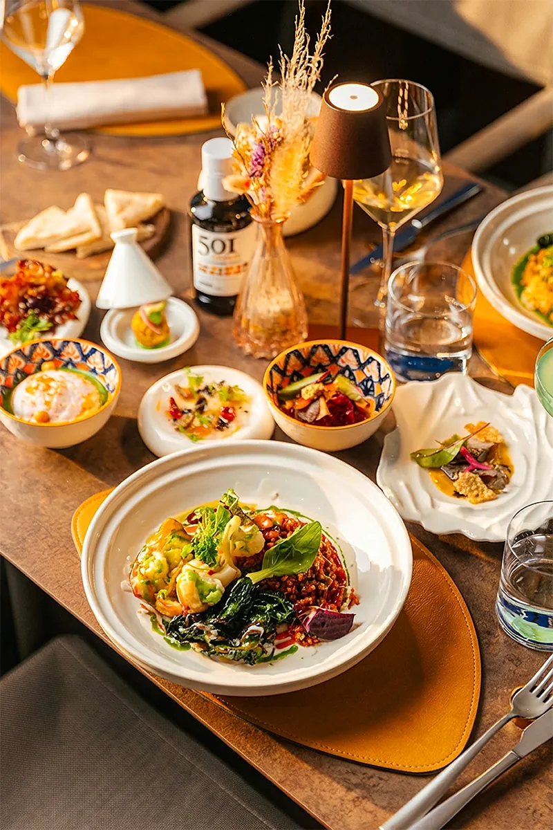Una tavola di Masa Roof Top con piatti da condividere, ciotole decorate e un calice di vino bianco